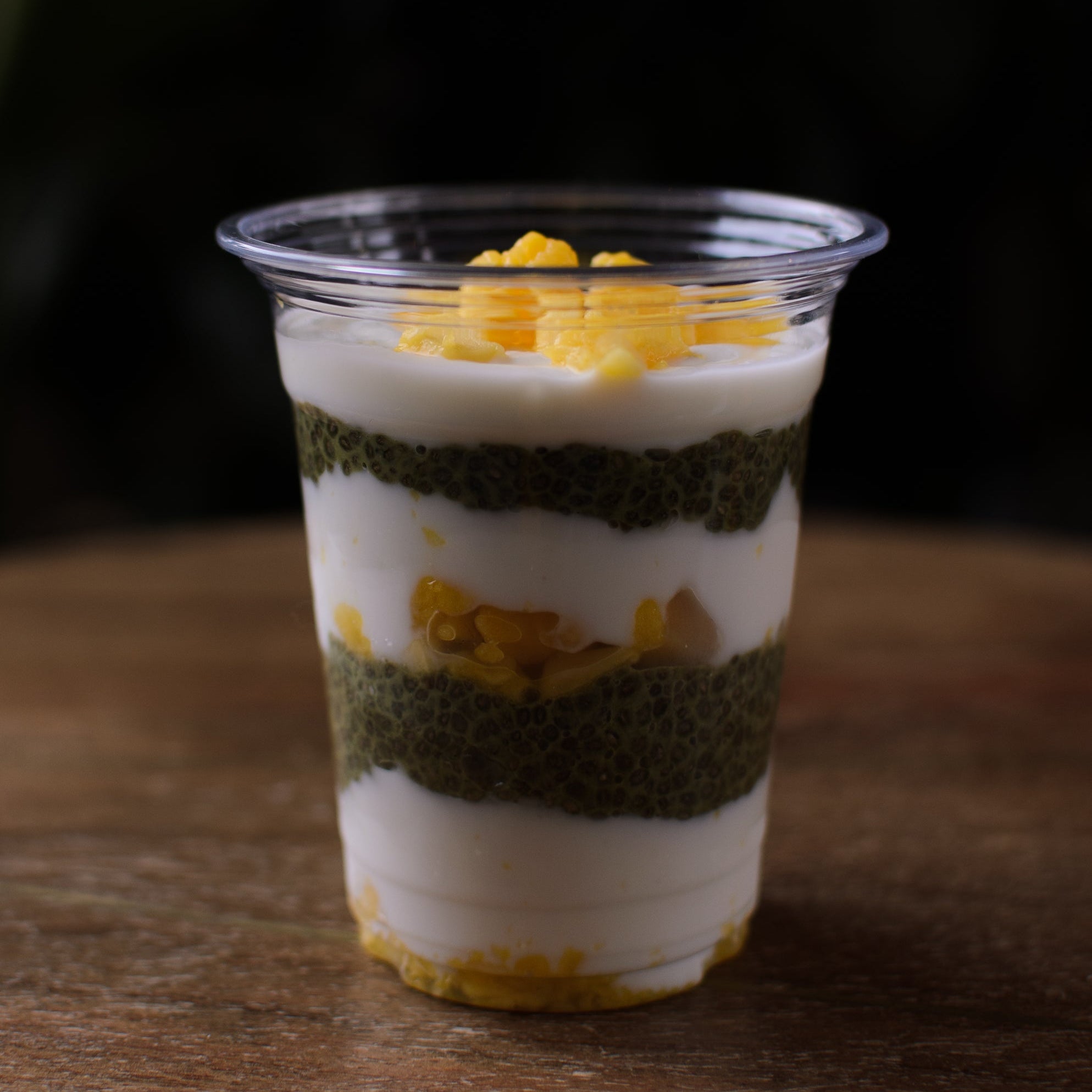 Layered dessert in a clear plastic cup on a wooden surface with a dark background