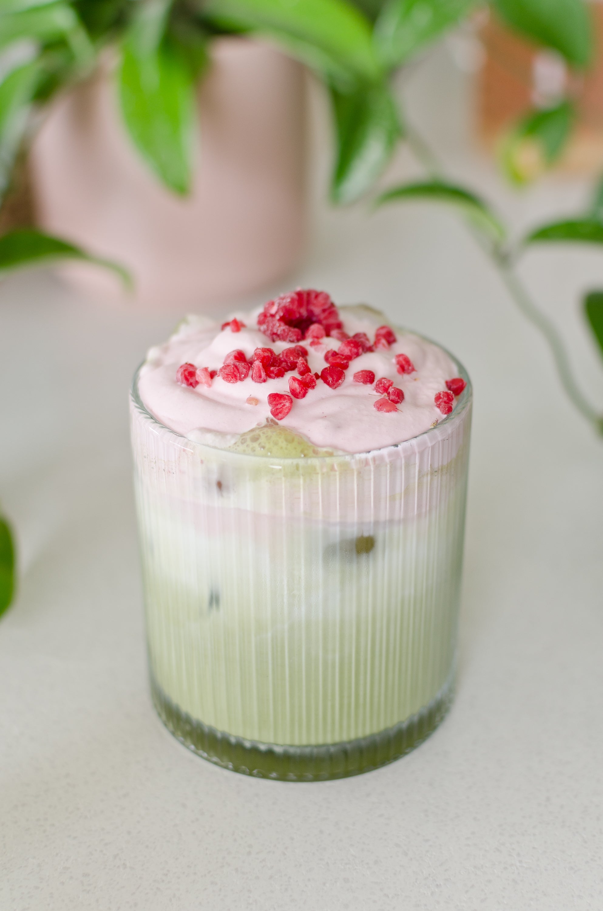 Iced matcha latte with pink cold foam and raspberries in a glass, set against a blurred green plant background.